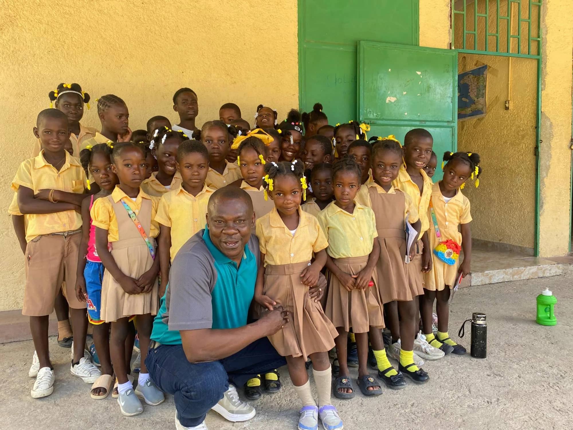 A staff member kneels with a class of students in yellow and tan uniforms outside their classroom door