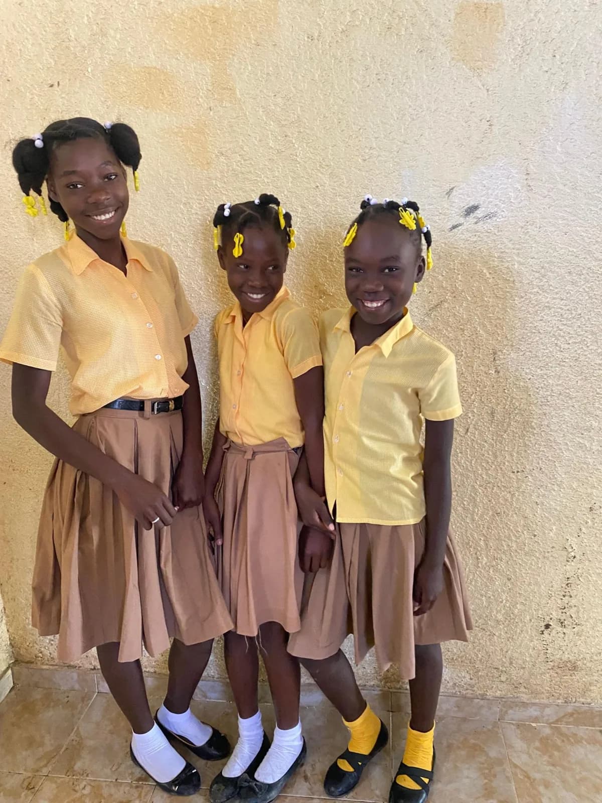 Three smiling girls in yellow and tan school uniforms stand together at an HEF school